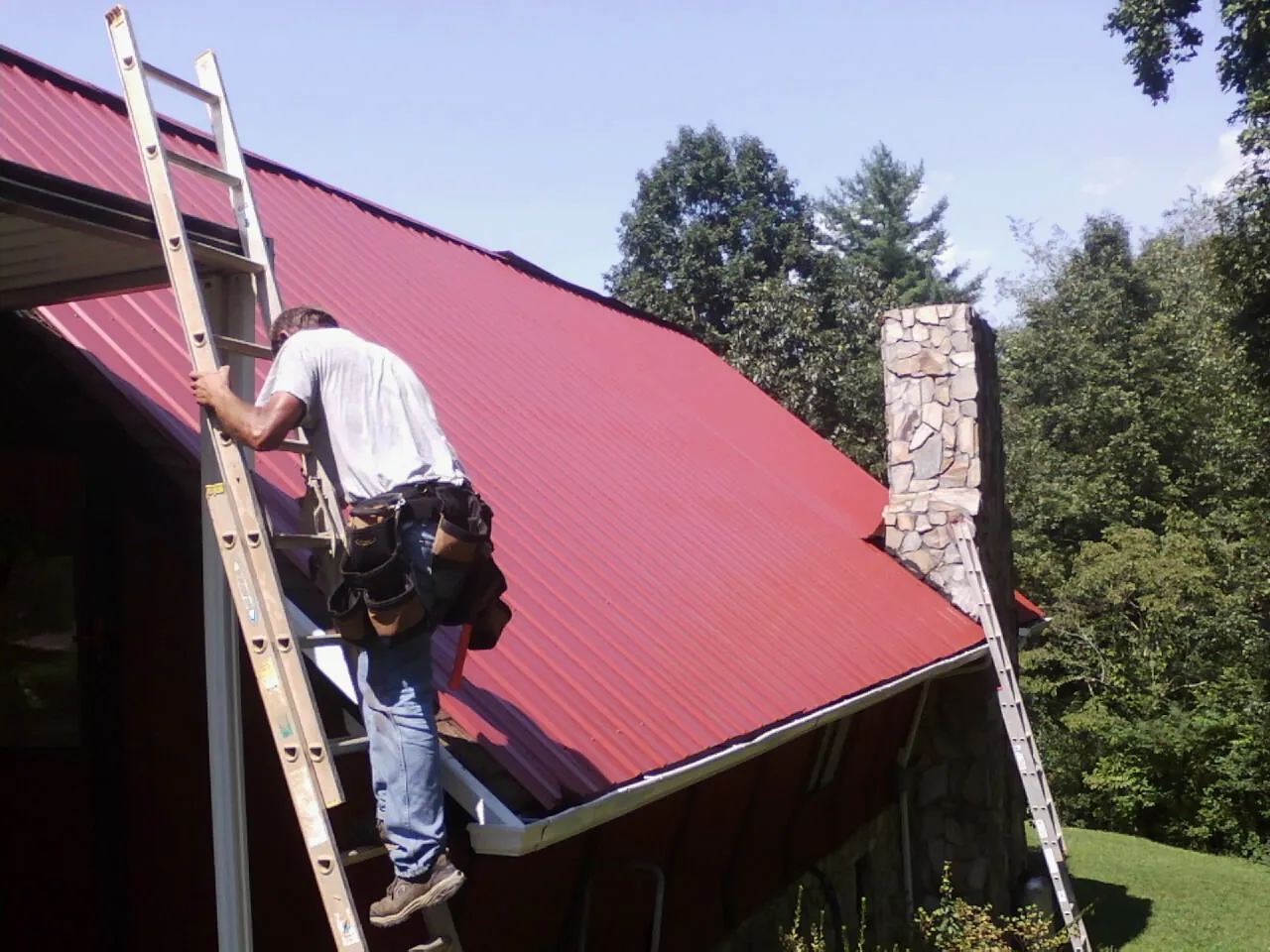 Roofer on a ladder beside a red metal roof and stone chimney — Soffit & Fascia Repair by Roofing Brothers in New Brunswick
